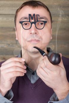 Young man with funny glasses on a stick and smoking pipe in hand, wooden background. Fun photo props and accessories for shoots