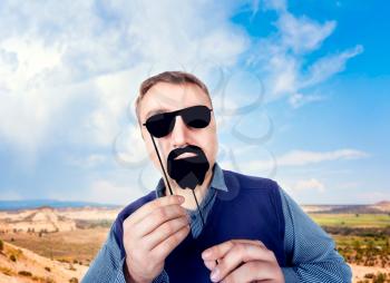 Young man with funny sunglasses and beard on a stick, mountain valley and blue sky on background. Fun photo props and accessories for shoots