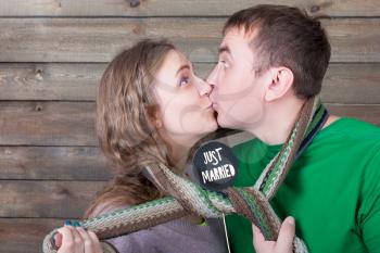 Love couple kisses wrapped in a scarf and shows funny icon on a stick with just married inscription, wooden background. Fun photo props and accessories for photo shoots
