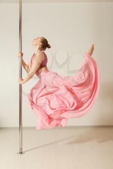 Sexy slim strip dancer exercising with pole in dance studio.  Attractive professional poledance girl posing in beautiful pink dress. 