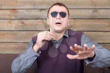 Playful man with funny sunglasses on a stick represents the blind person, wooden background. Fun photo props and accessories for shoots