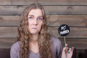 Young woman poses face and shows funny icon on a stick with just married inscription, wooden background. Fun photo props and accessories for photo shoots