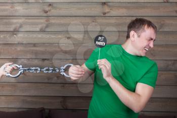 Young man handcuffed to marriage shows funny icon on a stick with  just married inscription, wooden background. Fun photo props and accessories for shoots