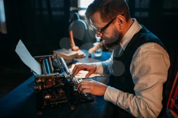 Bearded journalist in glasses typing on retro typewriter. Vintage lamp, feather, crystal decanter and books on background