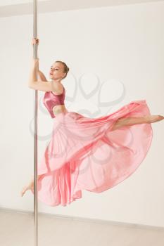 Sexy slim strip dancer exercising with pole in dance studio. Attractive professional poledance girl posing in beautiful pink dress.