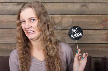 Young woman with disgusted face holding funny icon on a stick with just married inscription, wooden background. Fun photo props and accessories for shoots