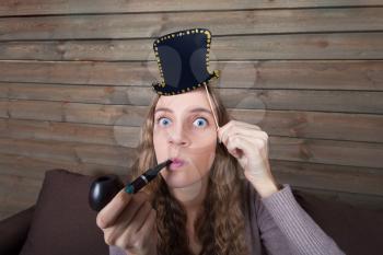Young woman with funny hat on a stick and smoking pipe in hand, wooden background. Fun photo props and accessories for shoots