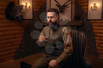 Hunter man in traditional vintage hunting clothing drink luxury alcohol after successful hunt. Fireplace, stuffed wild animals, bear skin and other trophies on background