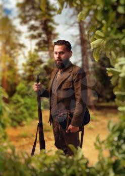 Portrait of breaded hunter man in traditional hunting clothing with old rifle, green forest on background. Hunt lifestyle