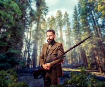 Portrait of breaded hunter man in vintage hunting clothing with old gun, green forest on background. Hunt lifestyle