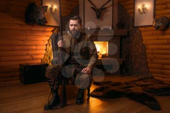 Vintage hunter man in traditional hunting clothing sitting in a chair with retro rifle against burning fireplace. Stuffed wild animals, bear skin and other trophies on background