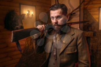 Respectable hunter man in vintage stylish hunting clothing with antique rifle against fireplace. Stuffed wild animals, bear skin and other trophies on background. Hunt lifestyle