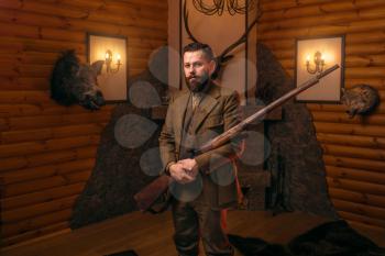 Hunter gentleman in traditional hunting clothing with old gun against fireplace. Stuffed wild animals, bear skin and other trophies on background. Old-fashioned style