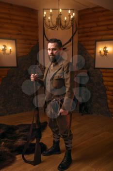Solid hunter man in old-fashioned hunting clothing with antique gun against fireplace. Stuffed wild animals, bear skin and other trophies on background. Hunt lifestyle