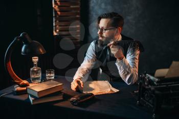 Thoughtful bearded writer in glasses smoking a pipe and look at the window. Retro typewriter, feather, crystal decanter, books and vintage lamp on the desk