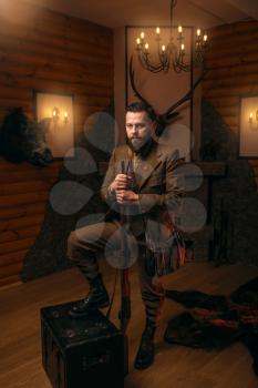 Hunter man with old gun in vintage traditional hunting clothing standing against antique chest. Fireplace, stuffed wild animals, bear skin and other trophies on background