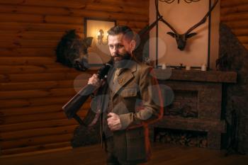 Respectable hunter man in vintage stylish hunting clothing with antique rifle against fireplace. Stuffed wild animals, bear skin and other trophies on background. Hunt lifestyle