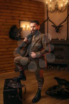 Respectable hunter man with old gun in retrro style traditional hunting clothing standing against antique chest. Retro rifle, vintage clothes. Room with fireplace, stuffed wild animals, bear skin and 