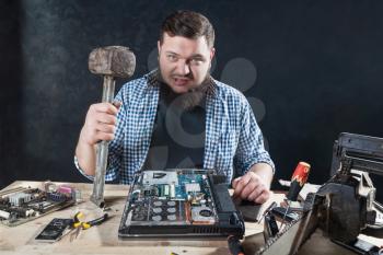 Service engineer with hammer, laptop and electronic components on the desk. Engineering humor