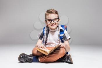Little boy with backpack reading a book in yoga pose, studio photo shoot. Young pupil in glasses with backpack and textbook. Child with schoolbag