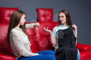 Young girl sitting on red leather couch and shows the costumes to the girlfriend. Leisure of happy girlfriends. Female friendship