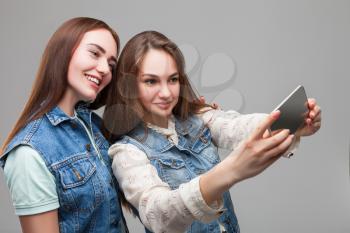 Two smiling girlfriends in denum jackets makes selfie on phone camera in studio. Female friendship. Leisure of happy girls 