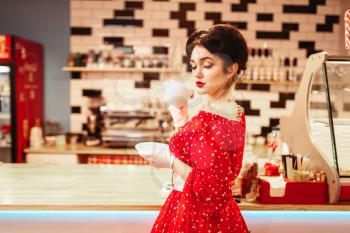 Glamour pin up girl with make-up drinks coffee in retro cafe, 50 american fashion. Red dress with polka dots, vintage style