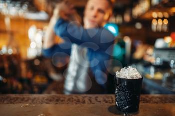 Black glass full of ice stands on bar counter, male barman with shaker on background. Barkeeper occupation, bartender job