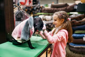Little girl chooses clothes for puppy in pet shop. Kid customer buying dogs overall in petshop, goods for domestic animals