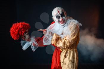 Mad bloody clown holds human skull in red wig, horror. Man with makeup in carnival costume, crazy maniac