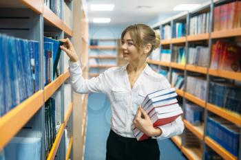 Pretty female person with stack of books between the shelves in university library. Young woman in reading room, knowledge depository
