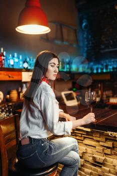 Young lady with glass of red wine sitting at wooden bar counter. Female customer leisures in pub