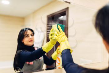 Housemaid hands in rubber gloves cleans the mirror with a cleaning spray, hotel bathroom interior on background. Professional housekeeping service, charwoman, sanitary processing