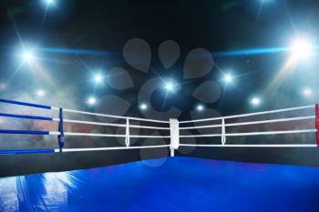 Empty boxing ring, blue flooring, view on corner with white ropes. Professional arena for sport competitions and fighting tournaments, nobody, bright spotlights on background