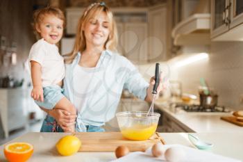 Young mother and her daughter mixing ingredients for cake in a bowl. Cute woman and little girl cooking on the kitchen, pastry preparation. Happy family prepares sweet desserts