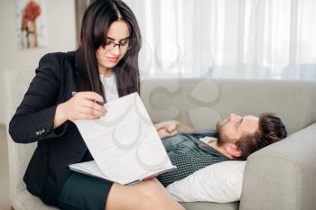 Stressed man lies on sofa at psychotherapist reception. Female doctor writes notes in notepad, professional psychology support