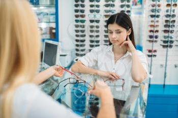 Female optician and woman chooses glasses frame in optics store. Selection of spectacles with professional optometrist
