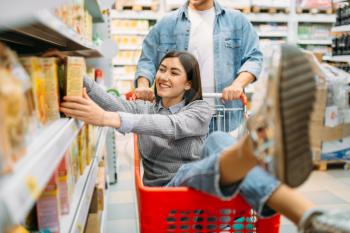 Man carries woman in the cart in supermarket. Family shopping. Playful couple in store