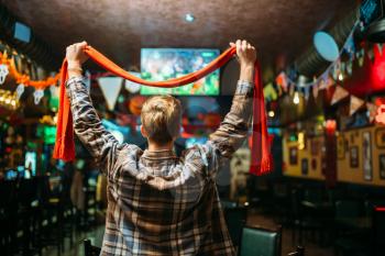 Football fan with scarf raises his hands up and watching the match of the favorite team in sports bar, back view. Tv broadcasting of the game in pub