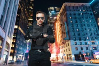 Police officer in sunglasses and black uniform with body armor, night city on background. Policeman in special ammunition