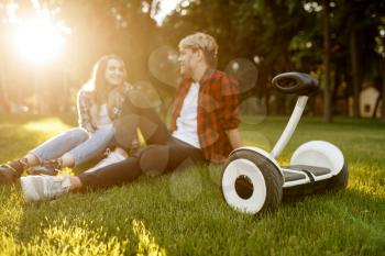 Young couple sitting on the grass near gyro board in summer park. Outdoor recreation with electric gyroboard. Eco transport with balance technology