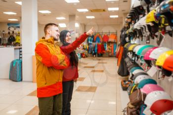 Young couple at the showcase choosing helmets for ski or snowboarding, side view, sports shop. Winter season extreme lifestyle, active leisure store, buyers looking on protect equipment