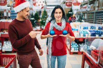 Funny couple chooses decorations in shop and jokes, woman puts christmas balls to her chest. December shopping of holiday goods