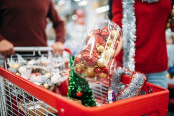 Young couple put in a cart Christmas decorations, purchasing in supermarket, family tradition. December shopping of holiday goods