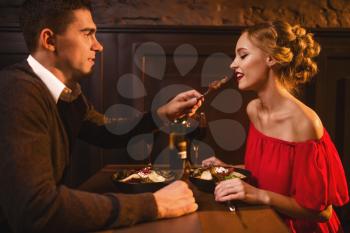 Young man meals elegant woman in red dress in restaurant. Beautiful love couple funs together, romantic evening 