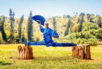 Female wushu master with fan sits on a twine, martial arts, green forest on background. Woman in blue cloth on outdoor fight training