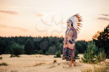 American Indian woman looks in a distance outdoors. Cherokee, Navajo culture. Headdress made of feathers of wild birds. Traditional costume