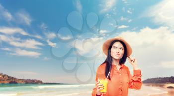 Happy woman in orange shirt and straw hat drinks freshly squeezedjuice, coastline. Young girl with yellow beverage, healthy lifestyle