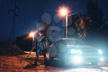 Serial maniac in leather coat and hat with bloody baseball bat wrapped in metal chain against black car with light at the night. Horror, bloody murderer, murder weapon