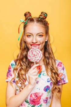 Beautiful young woman with playful look lick candy with tongue. Stylish girl with blonde curly hair. Portrait of attractive lady with big lollypop, yellow wall on background.